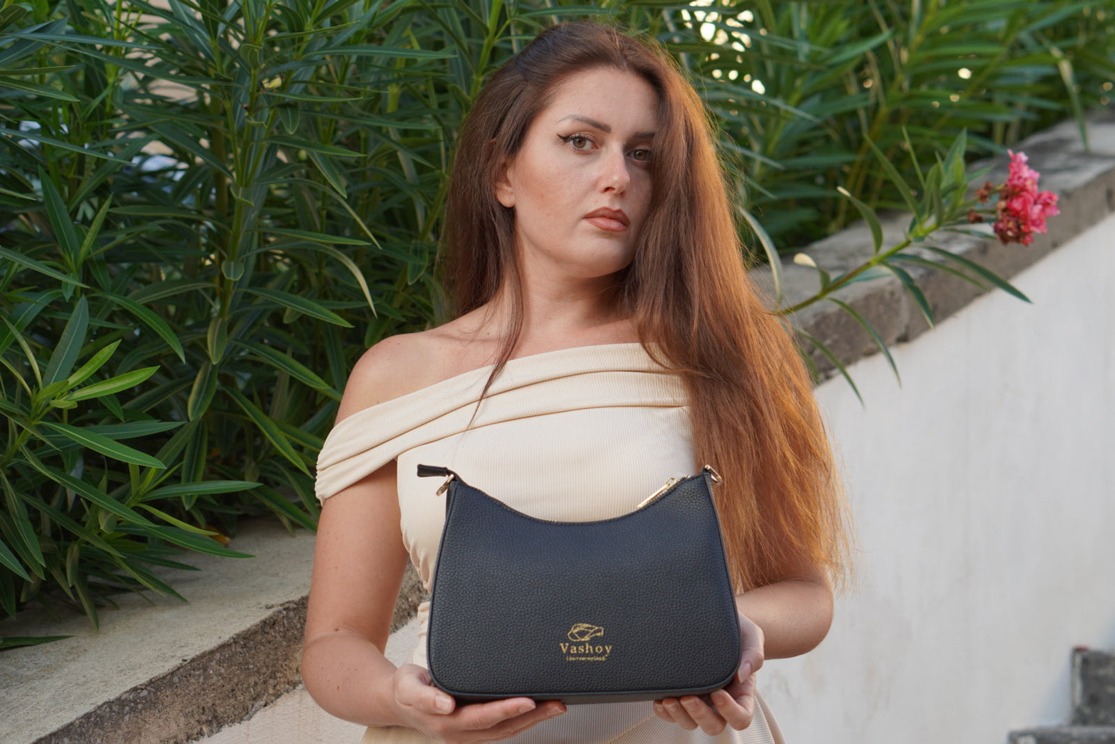 Woman in a cream off-the-shoulder top holding a black textured Vashoy bag with a gold logo outdoors beside green foliage.